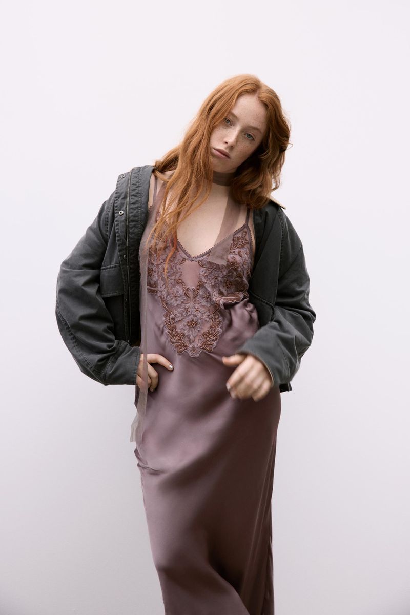 Adult woman with long red hair posing in a silky brown V-neck dress with lace details, wearing an olive jacket, standing indoors.