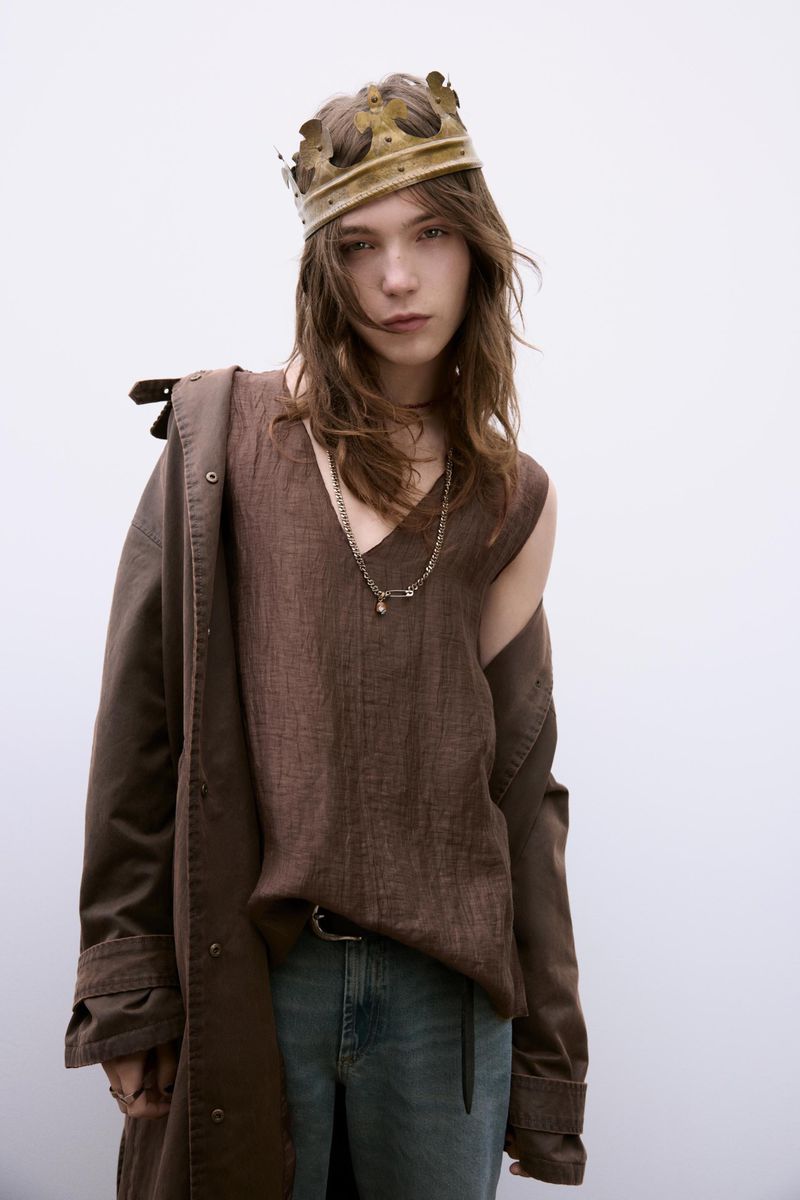 Person wearing a brown top, blue jeans, an overcoat, and a gold crown standing indoors in front of a white wall.