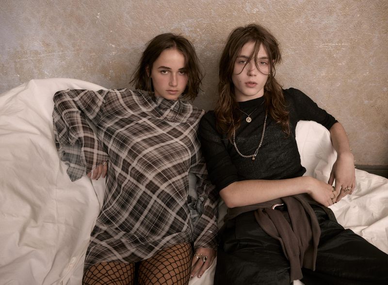 Two young adults in a casual indoor setting: one in a plaid dress with fishnet stockings, the other in dark clothing with long hair.