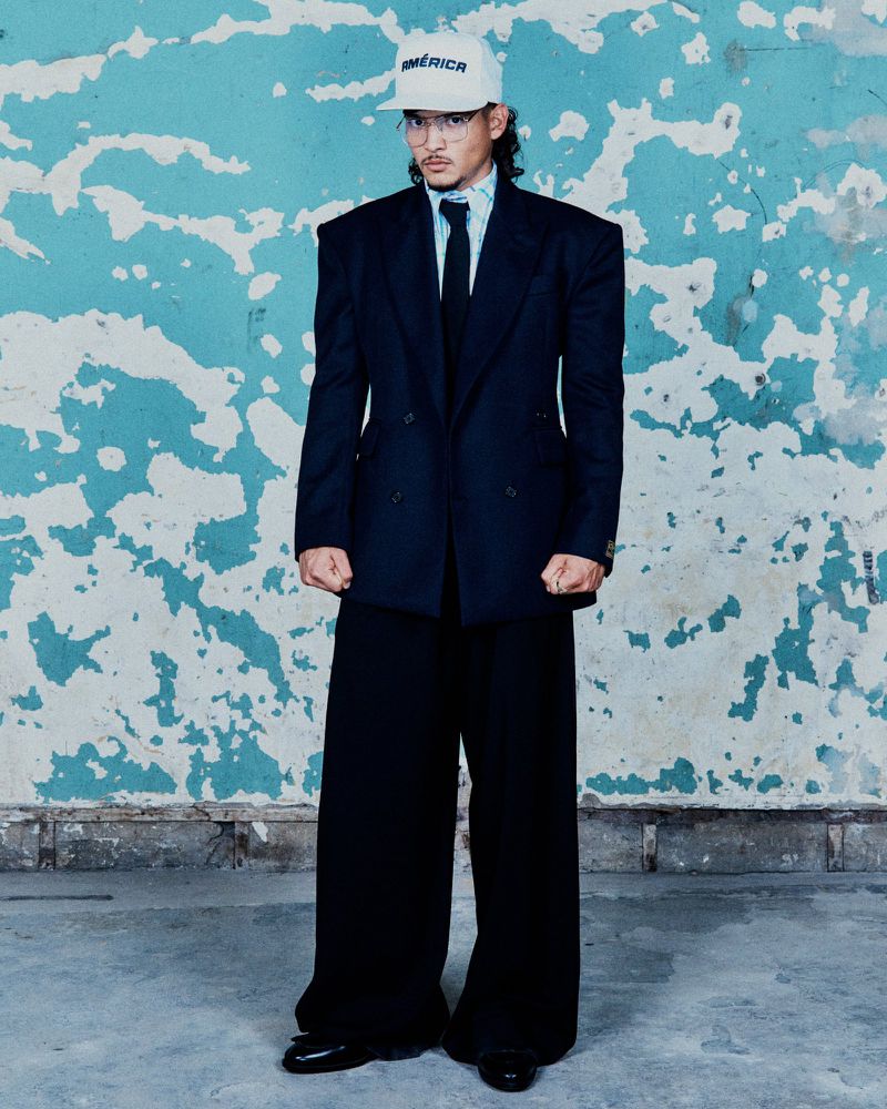 Adult male in oversized black suit and baseball cap with 'America' text, standing against a distressed blue wall, full shot.