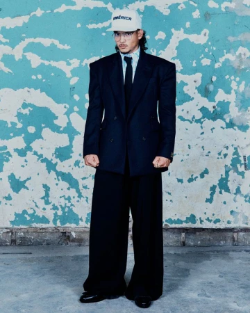Adult male in oversized black suit and baseball cap with 'America' text, standing against a distressed blue wall, full shot.