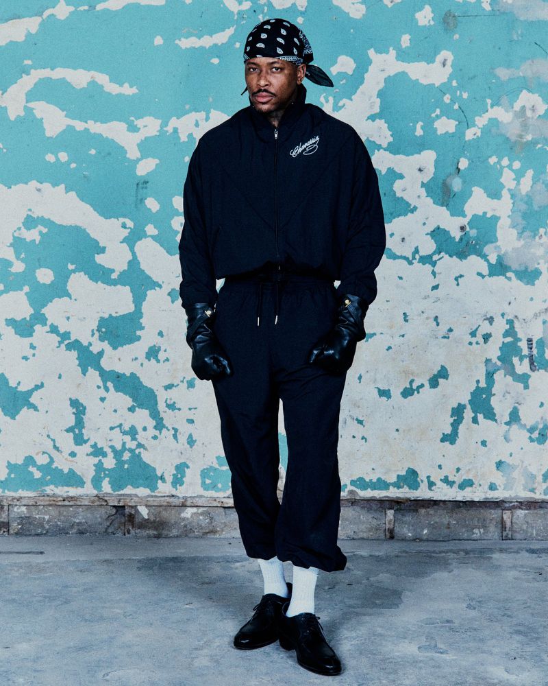 Adult male in a black outfit with black leather gloves poses in front of a distressed blue wall wearing a bandana and black dress shoes.