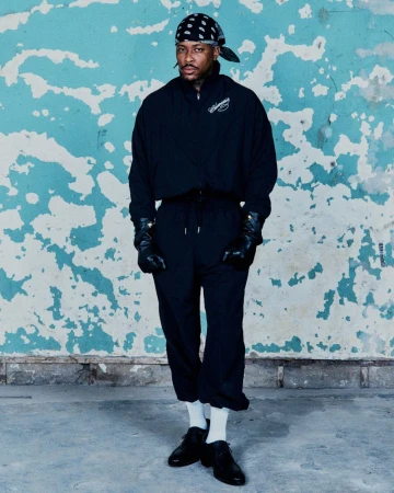 Adult male in a black outfit with black leather gloves poses in front of a distressed blue wall wearing a bandana and black dress shoes.