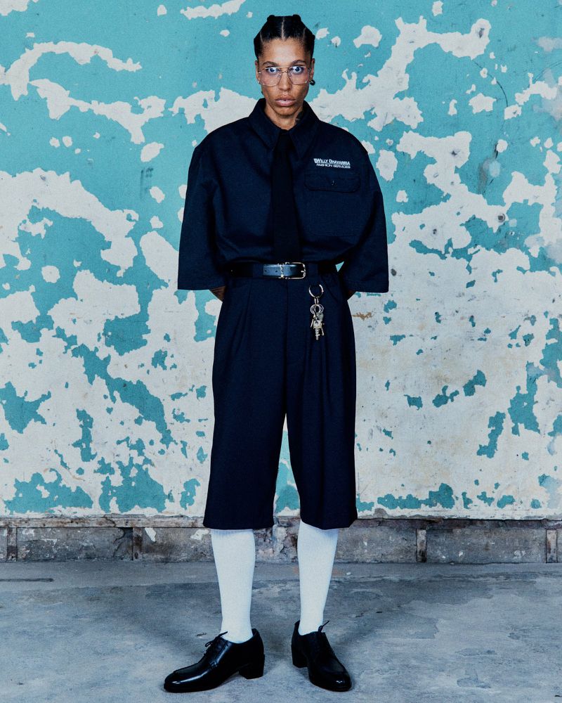 Model wearing an oversized black short-sleeve shirt, black tie, pleated knee-length shorts, and white socks against a peeling blue wall.
