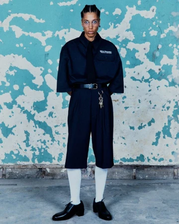 Model wearing an oversized black short-sleeve shirt, black tie, pleated knee-length shorts, and white socks against a peeling blue wall.