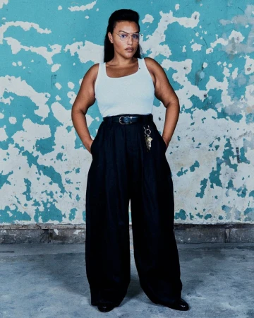Paloma Elsesser in a white tank top and oversized black wide-leg trousers standing before a distressed blue and white wall.