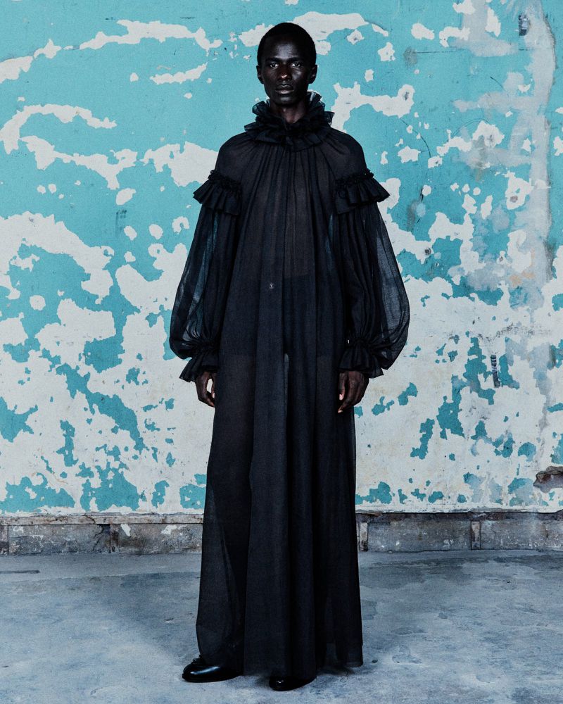 Fashionable adult male model in flowing black sheer gown with ruffles, standing indoors against a weathered wall with blue and white paint.