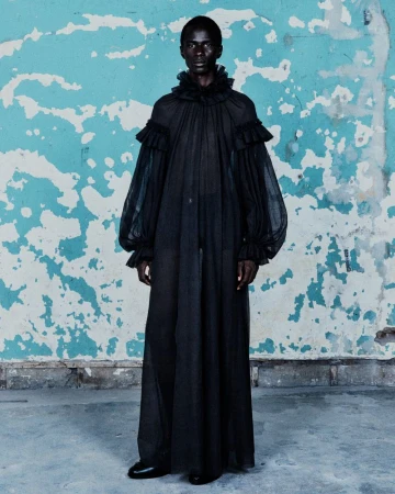Fashionable adult male model in flowing black sheer gown with ruffles, standing indoors against a weathered wall with blue and white paint.