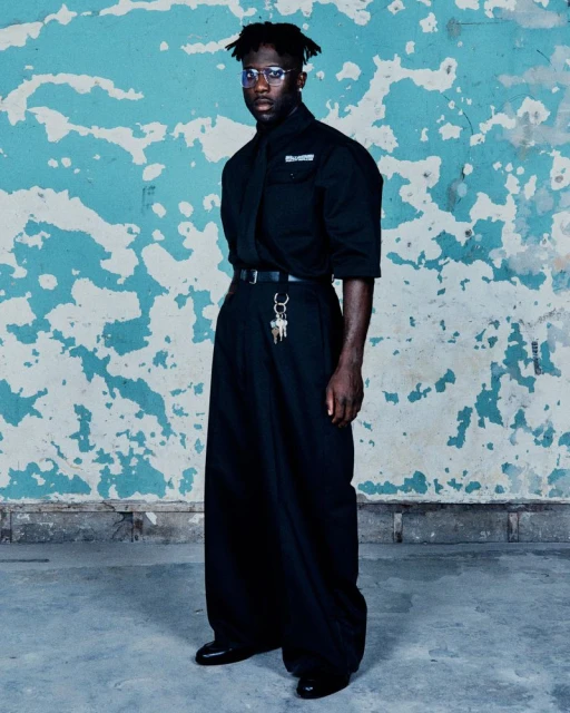 Full-length studio shot of a black man wearing an all black outfit against a textured blue wall. He is wearing glasses and has a serious expression.