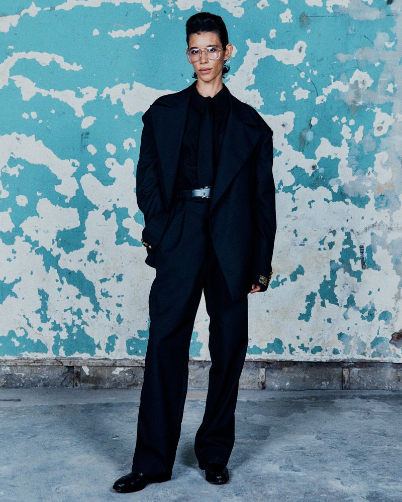 A person in a black suit with glasses stands against a blue and white distressed wall, creating a modern and stylish portrait.