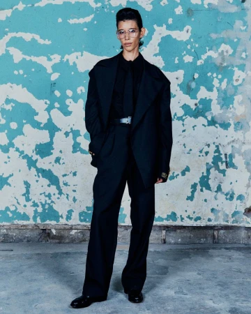 A person in a black suit with glasses stands against a blue and white distressed wall, creating a modern and stylish portrait.