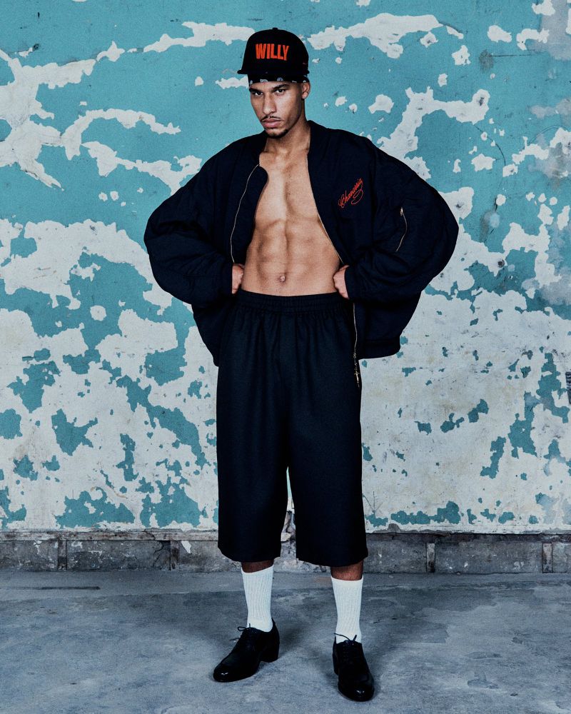 Man poses with open jacket, abs visible, black shorts, socks, black shoes against a distressed teal and white wall with black trim.