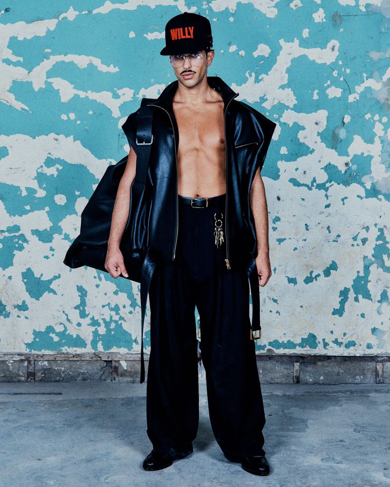 Male fashion model strikes a pose in a black leather vest, matching wide-leg pants, and a baseball cap against a textured blue wall.