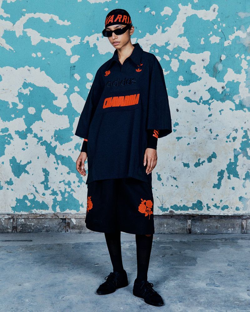 A stylish woman wearing an Adidas outfit with orange accents stands against a distressed blue wall in a fashion-forward pose.
