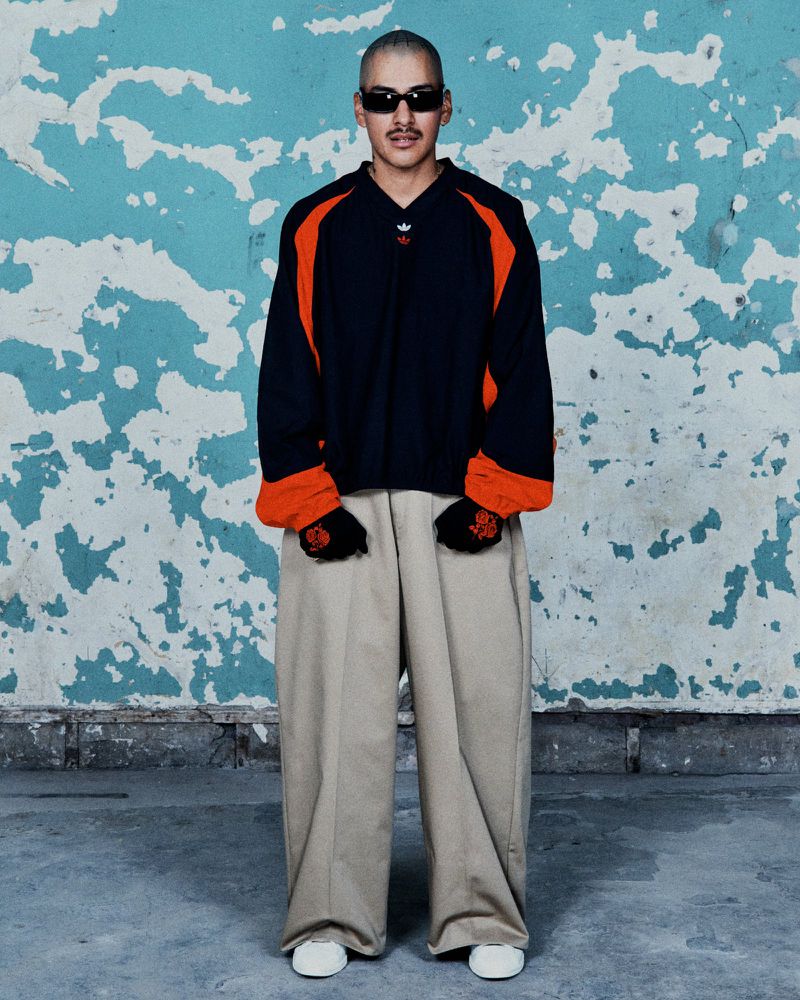 Man in streetwear fashion stands against a distressed blue wall, wearing a black and orange jacket, beige pants, white shoes, and sunglasses.
