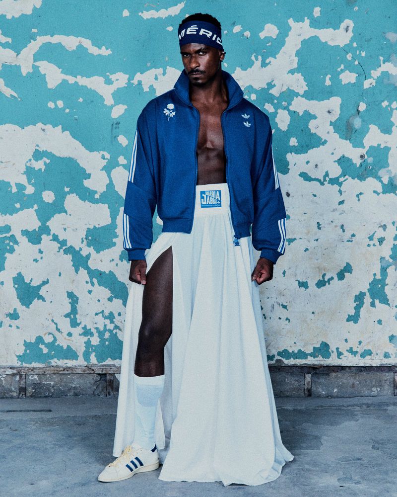 Athletic African American man poses in an Adidas jacket, skirt, and headband against a distressed turquoise wall indoors, showcasing unique style.
