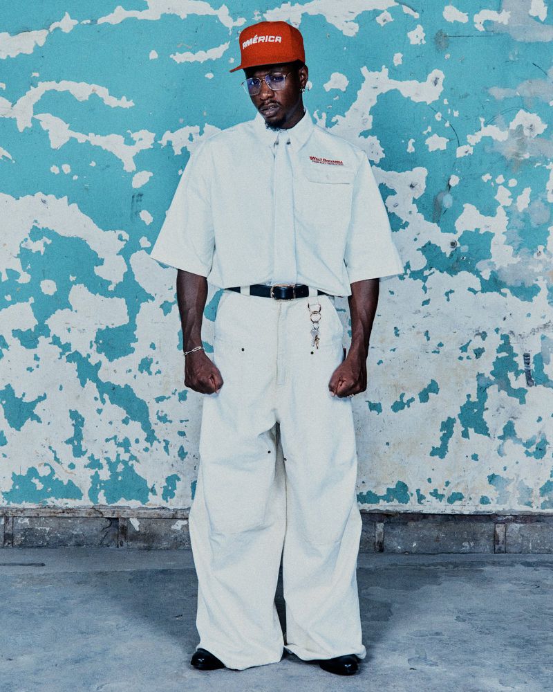 A man in a red cap and white outfit stands against a weathered blue and white wall, full-length shot with casual style and urban background.