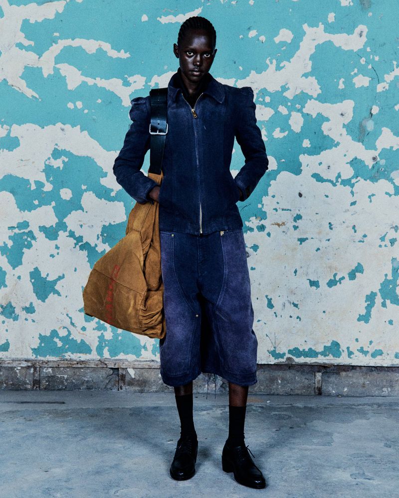 A dark-skinned adult stands in a studio wearing a navy jacket, culottes, black socks, and dress shoes, with a large tan duffle bag on shoulder.