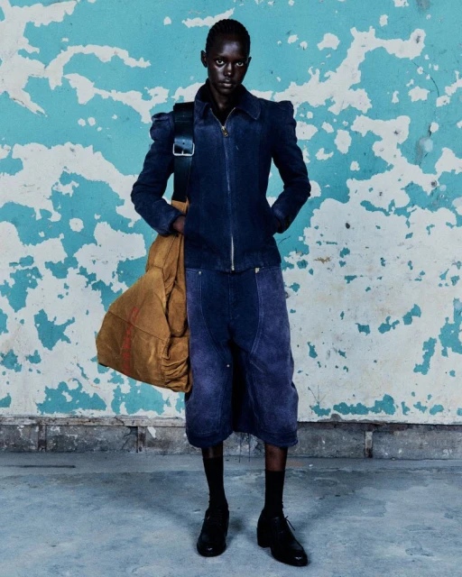 A dark-skinned adult stands in a studio wearing a navy jacket, culottes, black socks, and dress shoes, with a large tan duffle bag on shoulder.