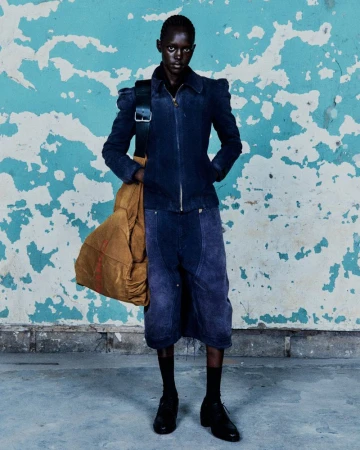 A dark-skinned adult stands in a studio wearing a navy jacket, culottes, black socks, and dress shoes, with a large tan duffle bag on shoulder.