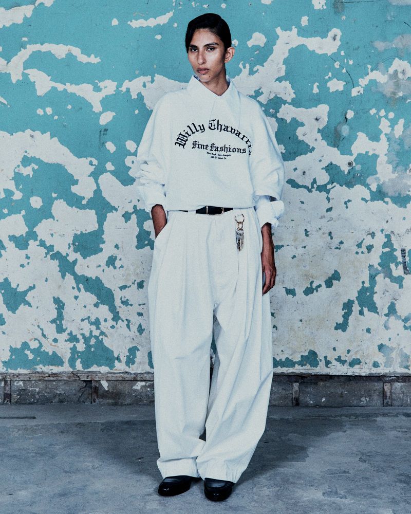 A model poses indoors against a distressed blue wall wearing a white sweatshirt, oversized white pants, and black shoes in a fashion shot.