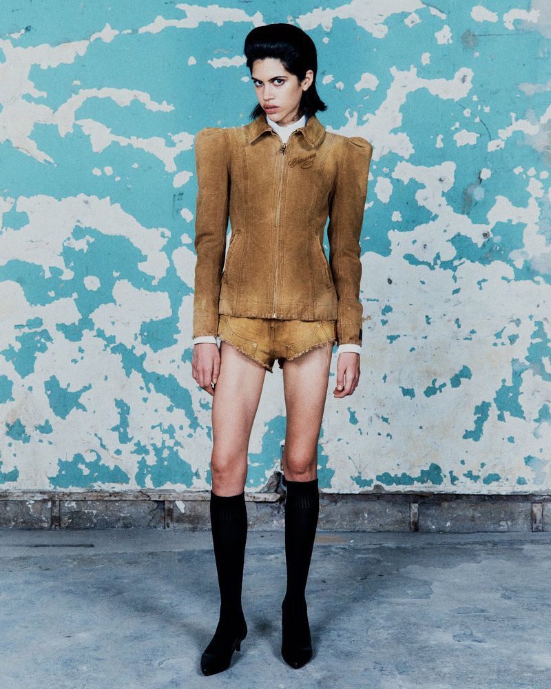 A full shot of a slim, fair-skinned model posing in a tan jacket and shorts in front of a worn, light-blue wall, wearing black socks and heels.