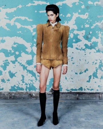 A full shot of a slim, fair-skinned model posing in a tan jacket and shorts in front of a worn, light-blue wall, wearing black socks and heels.