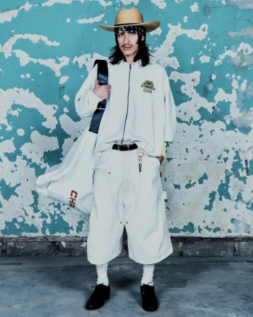 Model wearing a straw cowboy hat, oversized white zip-up top, and baggy knee-length shorts against a distressed turquoise wall.