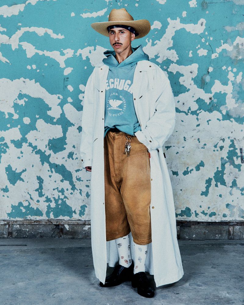 Stylish adult male in blue hoodie, tan pants, straw cowboy hat and black shoes, posing in front of a blue textured wall.