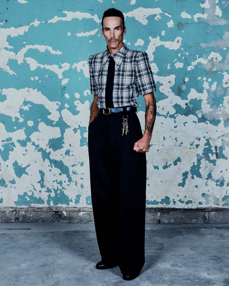 Man in oversized black pants and plaid shirt stands against an old turquoise wall with faded white patterns, accessorized with tie and keys.