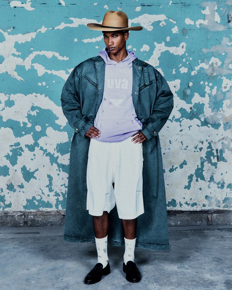 An African American man wearing a denim jacket, hoodie, shorts, cowboy hat and loafers poses against a distressed teal wall.