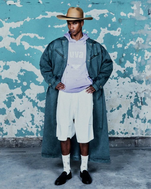 An African American man wearing a denim jacket, hoodie, shorts, cowboy hat and loafers poses against a distressed teal wall.