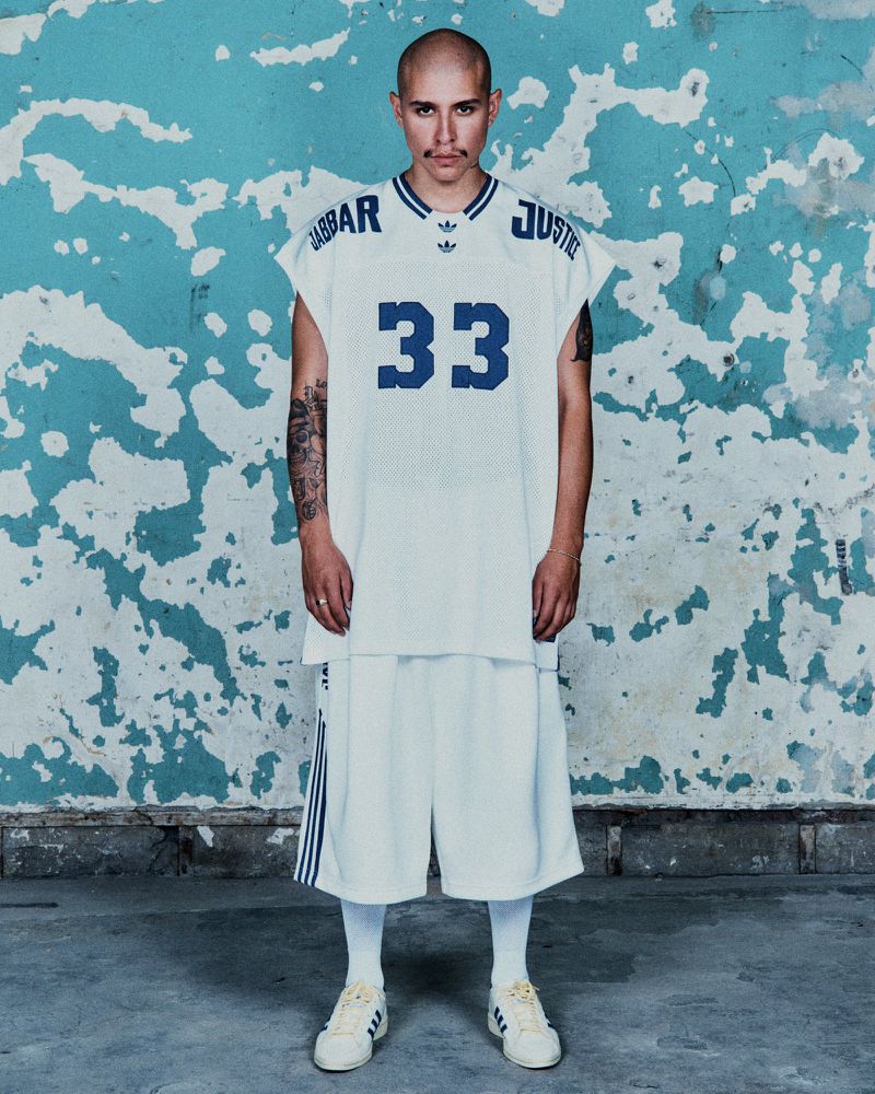 Young adult male poses in a vintage-style basketball jersey and shorts with iconic Adidas Superstar shoes against a distressed wall.