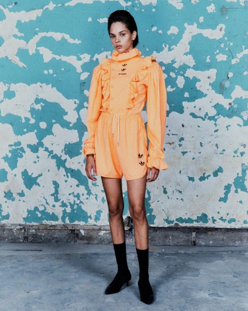 A woman in a peach Adidas jumpsuit with long sleeves and ruffled detailing poses in front of a weathered blue and white wall indoors.