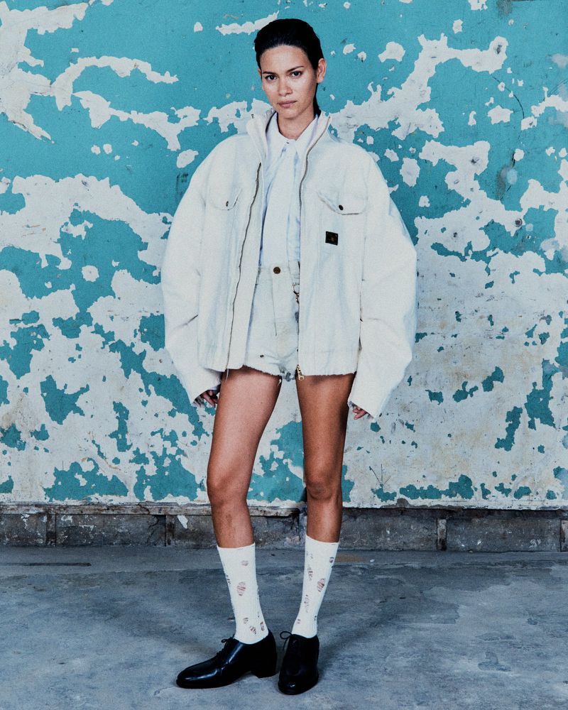 A stylish adult female posing in a white jacket and shorts against a distressed blue wall, complemented by socks and black dress shoes.