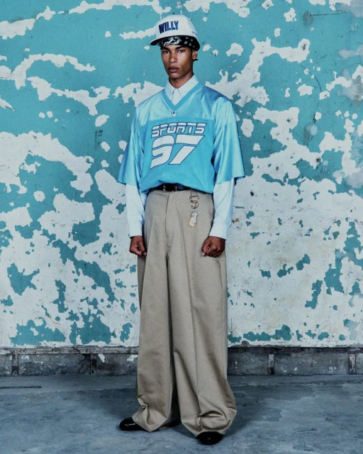 Young adult man posing in sports jersey and oversized pants against weathered wall. Casual fashion, urban style, cool expression.