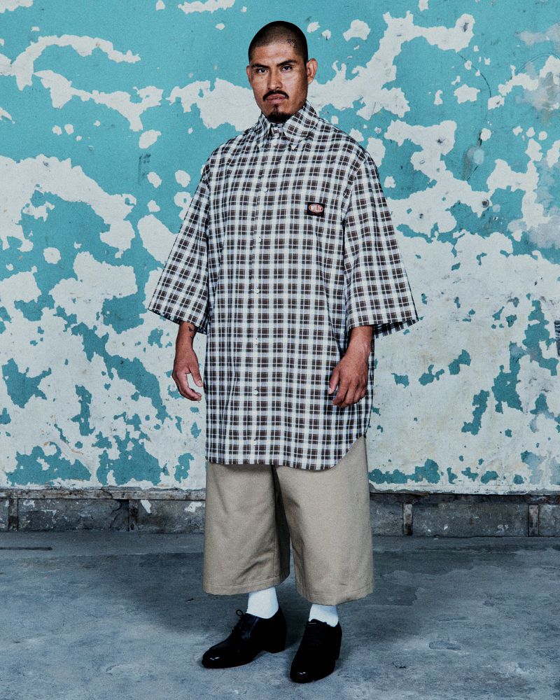 Adult man poses in a plaid, short-sleeved shirt and tan shorts against a distressed aqua wall, wearing black shoes and white socks.