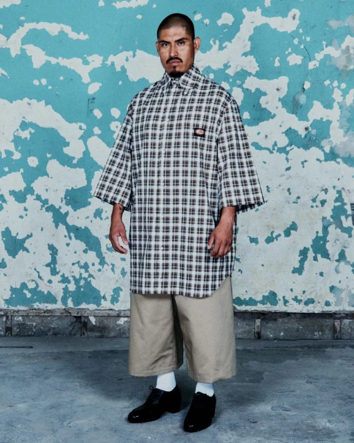 Adult man poses in a plaid, short-sleeved shirt and tan shorts against a distressed aqua wall, wearing black shoes and white socks.