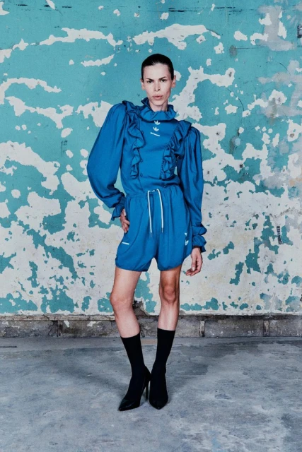 A slender adult female poses in a blue ruffled top and shorts with socks and heels against a teal and white textured wall indoors.