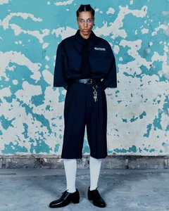 Model wearing an oversized black short-sleeve shirt, black tie, pleated knee-length shorts, and white socks against a peeling blue wall.