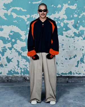 Model posing in an oversized black and orange long-sleeve top with wide-leg beige trousers against a distressed blue wall.