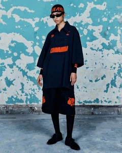 Model in oversized black Willy Chavarria polo, floral shorts, and headwrap standing against a peeling turquoise wall.