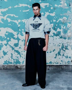 Man standing in grey printed sweatshirt and wide black trousers against a weathered teal and white wall.