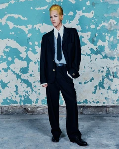 Man with yellow dyed hair posing in an oversized black suit, striped collared shirt, and necktie against a peeling turquoise wall.