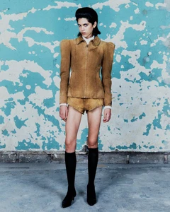 Model wearing a tan structured puff shoulder jacket with matching mini shorts, knee high black socks, and black heels.