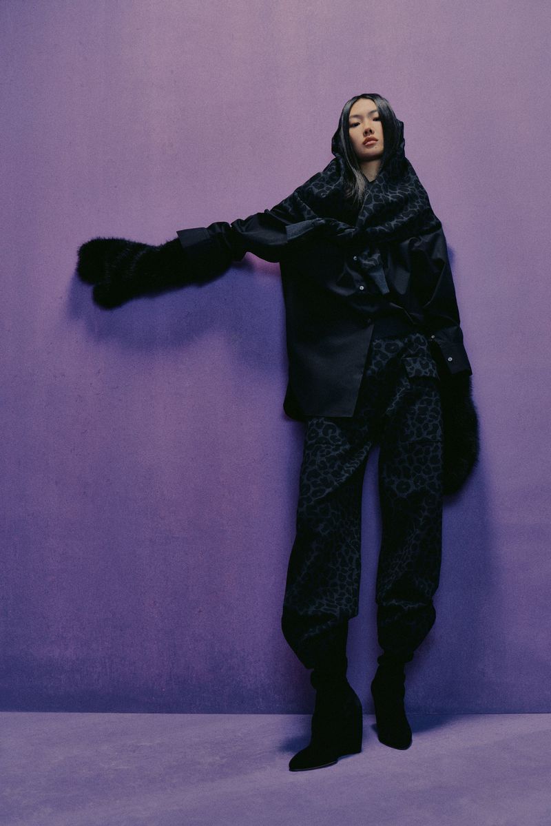 Adult female model in a black hooded jacket, black patterned pants and black boots, posing with an outstretched arm against a solid purple background.
