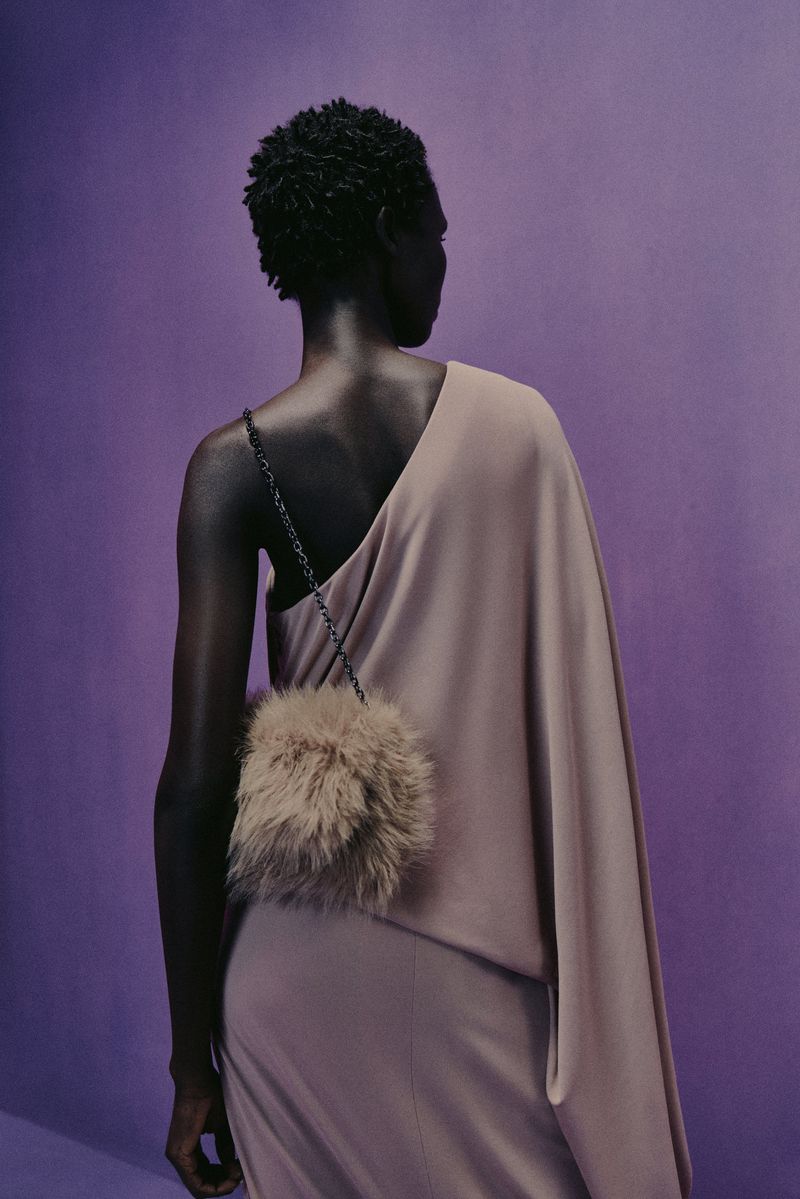 Back view of a Black woman in a taupe one-shoulder dress and fuzzy purse against a lilac studio backdrop in fashion photography.