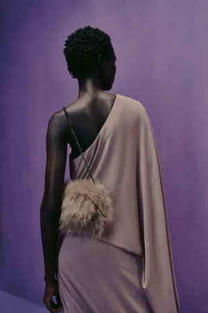 Back view of a Black woman in a taupe one-shoulder dress and fuzzy purse against a lilac studio backdrop in fashion photography.
