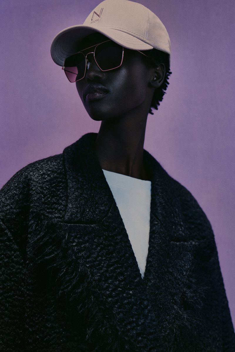 Close-up of a black woman wearing sunglasses and a baseball cap, posing against a purple backdrop in a dark textured coat.