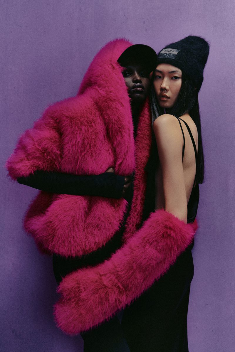 Two women pose against a lilac background, one in a bright pink fur coat and hat, the other in a black dress and beanie.